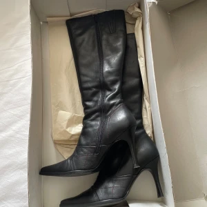 Svarta höga stövlar Buffalo London - Snygga svarta stövlar från Buffalo London i äkta skinn med hög klack och spetsig tå. Stövlarna har dragkedja på insidan och diskret logga upptill. Snygga sömdetaljer och mörkröd insida ger extra edge.😉