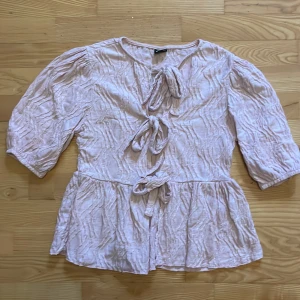 Ljusrosa blus med knyt och volang - Supersöt ljusrosa blus från Gina Tricot med tre knyt framtill och volang nertill. Blusen har ett krinklat tyg och trekvartsärmar med lätt puff. 