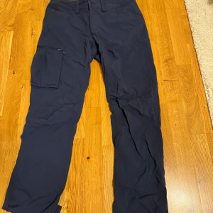 Mörkblå cargopants från Haglöfs - Snygga mörkblå cargopants från Haglöfs med praktisk benficka med dragkedja och diskret logga. Byxorna är raka i modellen och tillverkade i ett slitstarkt syntetmaterial, perfekta för uteliv eller chill vardagsstil.