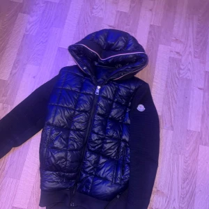 Svart dunväst med huva från Moncler - Snygg svart dunväst från Moncler med quiltad framsida och stor huva. Västen har dragkedja och stickade svarta ärmar med Moncler-logga på vänster arm. Perfekt för dig som vill ha en cool och sportig look. Säljer mellan 900-1100