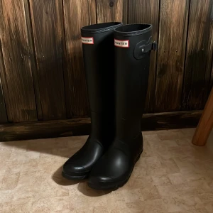 Svarta Hunter stövlar - Säljer ett par klassiska svarta Hunter stövlar i gummi med tillhörande gråa fleecefoder. Stövlarna är höga och har rund tå samt justerbar rem upptill. Perfekta för regniga dagar och håller fötterna varma med fodret. Stl 40/41. Har använt dom en gång, sen har dom stått, så är iprincip i nyskick! 🌸