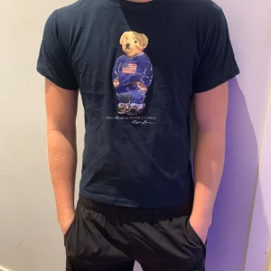 Mörkblå Polo Bear t-shirt Ralph Lauren - Snygg mörkblå t-shirt från Ralph Lauren med ikoniska Polo Bear-trycket på bröstet. Klassisk passform och rund halsringning, perfekt för en chill och stilren look. Tillverkad i mjuk bomull som känns skön hela dagen.