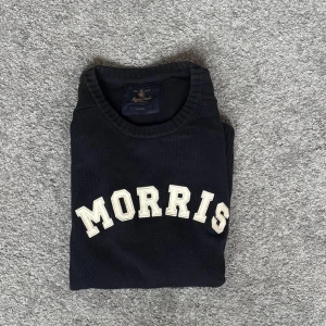 Mörkblå stickad tröja från Morris - Snygg mörkblå stickad tröja från Morris med stora vita bokstäver över bröstet. Klassisk rund halsringning och ribbade muddar vid ärmslut och nederkant. Perfekt för dig som gillar preppy stil och vill ha något bekvämt men ändå stilrent.