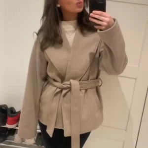 Beige omlottjacka med bälte - Snygg beige jacka med omlottmodell och brett bälte i midjan. Jackan har en avslappnad passform, stora slag och långa ärmar. Perfekt för lager-på-lager och ger en clean, stilren look. Materialet känns mjukt och har en ullig känsla.