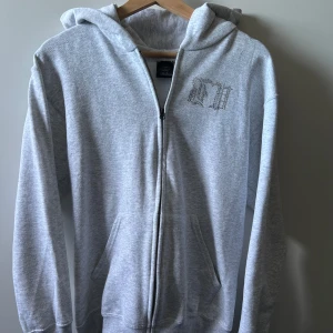 Grå Bvaldi hoodie med stenar - Grå hoodie med dragkedja och huva. Framtill finns ett diskret tryck på bröstet och på ryggen ett stort tryck. Mjuk bomullsblandning, kängurufickor och ribbade muddar. Perfekt för chill dagar eller streetstyle. Hoodien är aldrig använd så inte heller tvättad så alla stenar sitter kvar.