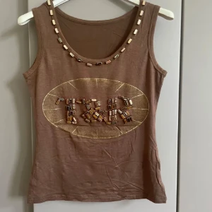 Brun linnetopp med pärldetaljer - Snygg brun linnetopp  med coola pärldetaljer runt halsringningen och på bröstet där loggan är markerad med pärlor i olika nyanser. Toppen är ärmlös och har en rund hals. Perfekt för dig som gillar unika detaljer och en avslappnad stil.