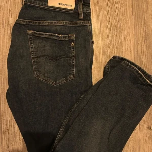 Replay mörkblå jeansbyxor - Snygga mörkblå jeans från Replay med klassisk femficksdesign och diskreta slitningar. Jeansen har raka ben och tydliga kontrastsömmar, samt Replay-logga på bakfickan och vid myntfickan. Perfekta för en avslappnad och stilren look.