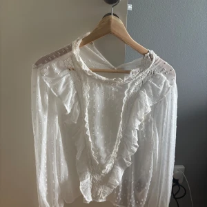 Vit blus med volang och spets från ONLY - Superfin vit blus från ONLY med volangdetaljer och spets längs v-ringningen. Blusen har långärmade transparenta ärmar och ett diskret prickigt mönster i tyget. Perfekt för dig som gillar romantisk och feminin stil. Blusen är även helt oanvänd!