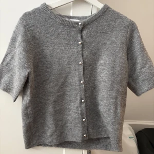 Grå stickad kofta  - Kortärmad grå stickad kofta med rund hals och snygga silvriga knappar framtill. Perfekt att slänga över en topp för en chill och stilren look. Mjuk och skön känsla tack vare det stickade materialet. Aldrig använd.
