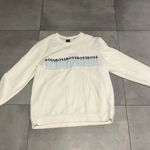 Vit sweatshirt från BOSS i XL - Vit sweatshirt från BOSS i storlek XL med tryckt BOSS-logga i flera rader över bröstet. Tröjan har rund hals, långa ärmar och ribbade muddar. Perfekt för en clean och stilren look. Materialet känns mjukt och skönt, troligtvis bomull.