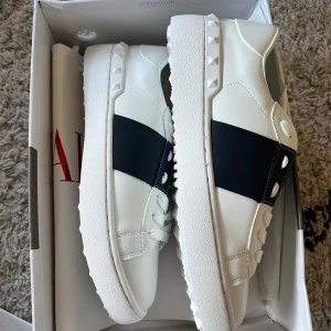 Valentino Garavani Open sneakers vit/svart - Snygga Valentino Garavani Open sneakers i vitt skinn med bred svart rem över mitten och ikoniska nitar på sulan. Klassisk rund tå, vita snören och guldfärgad logga på plösen. tyvärr för små
