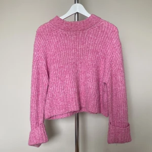 Rosa stickad tröja med uppvikta ärmar - Mysig rosa stickad tröja med grov ribbad struktur och uppvikta långa ärmar. Tröjan har en lite boxig, croppad passform och högre krage som ger en trendig look. Perfekt för kyliga dagar när du vill vara både bekväm och snygg.