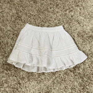 Vit volangkjol från Gina Tricot - Supersöt vit kortkjol från Gina Tricot med volangkant och smock i midjan. Kjolen har broderade detaljer och tunna spetsband som ger en romantisk vibe. Perfekt för sommarens alla häng! Materialet är mjukt och lätt bomull.