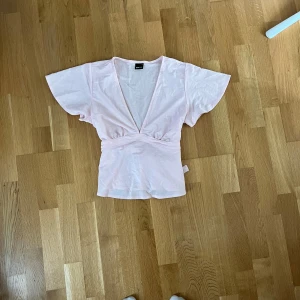 Ljusrosa blus från Gina Tricot - Supersöt ljusrosa blus från Gina Tricot med korta, vida ärmar och knytband i midjan. Toppen har en djup v-ringning och är gjord i mjuk bomull. Använd 1 gång💗
