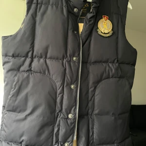 Svart dunväst från Morris Ivy League - Säljer en svart dunväst från Morris Ivy League med broderad patch på bröstet. Västen har två stora fickor framtill, dragkedja och tryckknappar. Insidan är fodrad i beige och västen har en klassisk, quiltad look. Perfekt för lager-på-lager-stil.