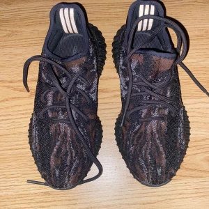 Adidas Yeezy Boost 350 V2 mörka sneakers - Säljer ett par Adidas Yeezy Boost 350 V2 med svart och brun stickad ovandel, svarta snören och en chunky svart sula. Skorna har de klassiska tre ränderna på hälen och är tillverkade i syntetmaterial med en unik mönstrad design.