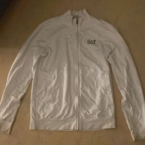 Vit zipjacka från EA7 Emporio Armani - Snygg vit jacka från EA7 Emporio Armani med dragkedja hela vägen och diskret logga på bröstet. Jackan har lång ärm, ståkrage och två fickor framtill. Perfekt för en clean och sportig look. Pris är förhandlingsbart 