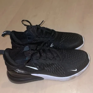 Air Max 270 Nike - Nike Air 270 sneakers i svart med vit sula och diskreta vita detaljer. Skorna har mesh-överdel för bra ventilation, snörning och en synlig Air-enhet i hälen som ger extra komfort. Perfekt för dig som gillar sportig och modern stil. Helt nya och oanvända 