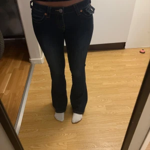 True Religion Joey Low Rise Flare Jeans - Säljer ett par mörkblå True Religion Joey jeans med låg midja och flare/bootcut. Klassiska kontrastsömmar, snygga fickdetaljer bak och coola knappar. Jeansen är i stretchigt denim och har en riktigt schysst passform som framhäver figuren.