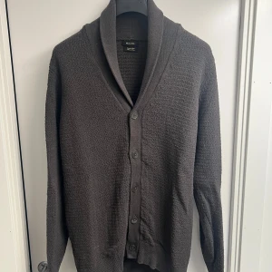 Mörkgrå kofta från Massimo Dutti - Säljer en stilren mörkgrå kofta från Massimo Dutti med sjalkrage och knäppning framtill. Koftan är stickad och har långa ärmar samt ribbade muddar. Perfekt för lager-på-lager och passar till många olika outfits. Köpt på Massimo Dutti i Paris.