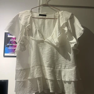 Vit volangblus med v-ringning - Superfin vit blus med v-ringning och stora volangdetaljer längs ärmar och nederkant. Luftig och flowy passform, perfekt för dig som gillar romantisk och trendig stil. Tunt, lätt material som känns skönt mot huden. Helt oanvänd 