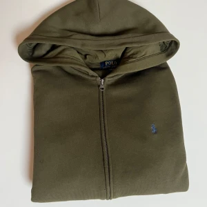 Olivgrön hoodie från Polo Ralph Lauren - Snygg olivgrön hoodie från Polo Ralph Lauren med dragkedja och huva. Klassisk broderad logga i blått på bröstet. Tillverkad i mjuk bomullsmix, perfekt för chill dagar. Enkel och stilren design som funkar till det mesta.