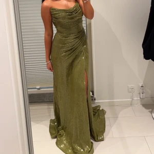 Grön glittrig långklänning med slits - Säljer en snygg grön långklänning med glittrigt tyg och draperad överdel. Klänningen är axelbandslös och har en slits framtill. Perfekt för fest och bröllop.