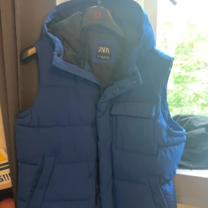 Blå dunväst från Zara - Snygg blå dunväst från Zara med hög krage och flera praktiska fickor framtill. Västen är quiltad och har en sportig look, perfekt att slänga över en hoodie eller tröja. Stängs med både dragkedja och knappar.