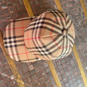 Burberry rutig keps i beige och svart - Säljer en snygg Burberry keps med klassiskt rutigt mönster i beige, svart, vitt och röda linjer. Kepsen har justerbar snapback och en svart patch med 'Burberry London England' bak. Perfekt för dig som vill ha en ikonisk streetstyle-accessoar.