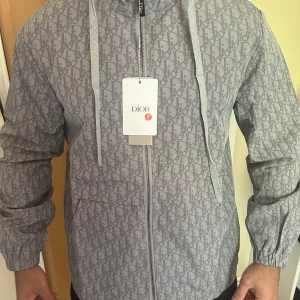 Grå vindjacka  - Snygg grå vindjacka med ikoniskt all-over logomönster. Jackan har huva med snörning, dragkedja framtill och elastiska muddar vid ärmslut. Perfekt för dig som vill sticka ut med en exklusiv streetstyle-look.