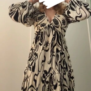 Snygg klänning - Säljer en luftig långklänning i vitt med svart abstrakt mönster. Klänningen har v-ringning, långa vida ärmar och plisserad struktur. Perfekt för dig som gillar en bohemisk och trendig stil. 
