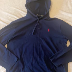 Marinblå hoodie från Polo Ralph Lauren - Säljer en marinblå hoodie från Polo Ralph Lauren med klassisk röd broderad logga på bröstet. Tröjan har huva med snörning och är tillverkad i mjuk bomull. Perfekt för dig som gillar stilrena och bekväma plagg. Ganska använd men inga defekter. Nypris 1500