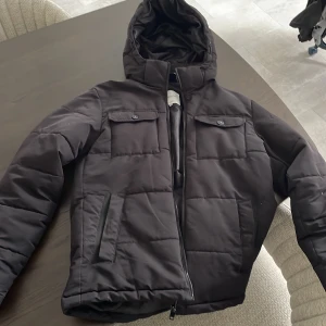 Svart pufferjacka från Jack & Jones XS - Säljer en svart pufferjacka från Jack & Jones i storlek XS. Jackan har huva, två bröstfickor med knappar och två sidofickor med dragkedja. Perfekt för kalla dagar med sin quiltade design och stilrena look.