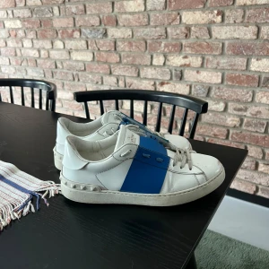 Valentino Garavani Open sneakers vit/blå - Snygga Valentino Garavani Open sneakers i vitt skinn med bred blå rem över vristen och coola nitar på hälen. Klassisk rund tå, platt sula och vita snören. Kommer med originalkartong, dustbag och extra skosnören. Perfekta för dig som gillar exklusiva streetwear-vibbar. 