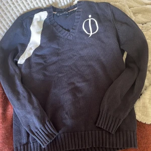 Mörkblå stickad tröja Oscar Jacobson - Säljer en mörkblå stickad tröja från Oscar Jacobson Sports med vit detalj på axeln och broderad logga på bröstet. Tröjan har v-ringning, långa ärmar och breda ribbade muddar vid ärmslut och nederkant. Perfekt för dig som gillar klassisk stil.