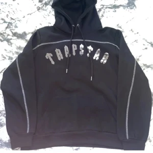 Svart Trapstar hoodie med snörning - Svart hoodie från Trapstar i storlek S med coolt tryck på bröstet och vita kontrastsömmar längs ärmar och axlar. Tröjan har huva med snörning och en stor magficka. Perfekt för dig som gillar streetwear och vill sticka ut. Tyvärr måste jag sälja den för den blev för liten, efterlängtad tröja som har vart använd 1-3 gånger, hör av er vid minsta fundering:)