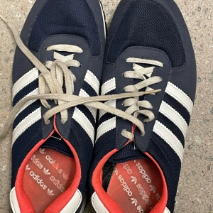 Adidas sneakers marinblå/vita strl 40⅔ - Snygga Adidas sneakers i marinblått med klassiska vita ränder och detaljer. Skorna har röd insida, vit häl och beige snörning. Ovandelen är i textil och mocka, med en slitstark gummisula. Perfekta för dig som gillar retrostil och streetwear.