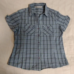 Blågrön rutig kortärmad y2k skjorta IMAGE - Kortärmad skjorta från IMAGE i blågrönt rutigt mönster. Skjortan har två bröstfickor med knappar, klassisk krage. Tillverkad i 100% bomull, perfekt för en avslappnad stil. Säljer en annan från samma märke i annat mönster också. Storlek 42 men passar fler storlekar, beroende på hur man vill att den ska sitta (se mått i den andra annonsen). Bara använd någon enstaka gång - i fint skick.