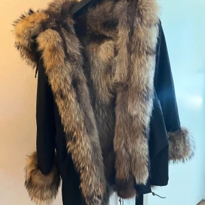 Svart parkajacka med stor päls - Parkajacka i svart med massiv brun päls runt huva, ärmslut och insida. Jackan har snörning i midjan och en oversized look. Pälsen är fluffig och ger jackan en riktigt lyxig vibe, perfekt för kalla dagar när du vill sticka ut.