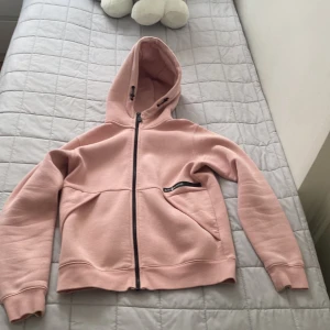 Ljusrosa hoodie från Sail Racing - Ljusrosa hoodie från Sail Racing med dragkedja framtill och huva med snörning. Tröjan har två fickor och svart logga på bröstet. Mjuk insida och ribbade muddar vid ärmslut och nederkant. Perfekt för chill dagar eller när du vill vara extra bekväm. 