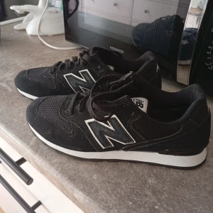 Svarta New Balance sneakers - Snygga svarta New Balance sneakers med klassisk vit sula och stort N på sidan. Ovandelen är i mocka och mesh för en sportig look. Skorna har snörning och en platt, mönstrad sula som ger bra grepp. Perfekta för dig som gillar stilrena och bekväma sneakers.