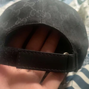 Svart keps från Gucci med GG-mönster - Snygg svart keps från Gucci med klassiskt GG-monogram över hela kepsen. Kepsen har justerbar rem bak i skinn och en ikonisk grön-röd rand på sidan. Materialet är kraftig bomullscanvas och detaljer i skinn. Perfekt för dig som vill sticka ut med lyxig streetstyle.