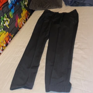 Svarta kostymbyxor med smal rand - Snygga svarta kostymbyxor med tunna vita ränder. Klassisk rak passform och diskret dragkedja framtill. Perfekta för dig som gillar stilrena och dressade vibes. Materialet känns slätt och har en lätt glans.