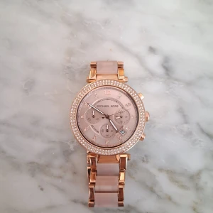 Michael Kors klocka - Snygg Michael Kors klocka i roséguld med glittrande stenar runt urtavlan. Klockan har länkarmband i metall och plast och datumvisning. Använd fåtal gånger och har ny bytt batteri💕 Omkrets 19,5cm💕