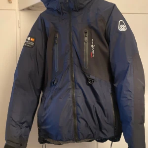 Sail Racing blå dunjacka M - Blå och svart dunjacka från Sail Racing med huva, flera dragkedjeförsedda fickor och coola loggor på ärmen och bröstet. Jackan har sportig look, justerbar nederkant och är perfekt för kalla dagar. Insidan är ljusgrå och materialet känns slitstarkt och vattenavvisande. Denna modell säljs inte längre vilket gör den unik.