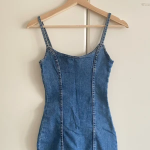 Blå jeansklänning med dragkedja - Säljer en snygg blå jeansklänning med smala axelband och figurnära passform. Klänningen har en cool dragkedja i ryggen och sömmar som ger en schysst siluett. Perfekt för dig som gillar denim och vill ha en enkel men trendig look.