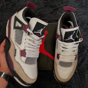 Nike Air Jordan 4 PSG sneakers - Nike Air Jordan 4 PSG sneakers i vitt, beige och vinrött med svarta detaljer. Skorna har meshpaneler, snörning och Paris Saint-Germain-logga på hälen. Tillverkade i skinn och mocka med platt sula och ikonisk Jumpman-logga på tungan.