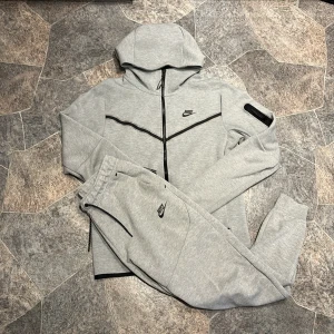 Grå Nike hoodie med dragkedja - Säljer en stilren ljusgrå hoodie från Nike med svart logga på bröstet och svarta detaljer. Tröjan har hel dragkedja, huva och långa ärmar. Perfekt för chill eller träning. Materialet är mjukt och känns skönt mot huden.