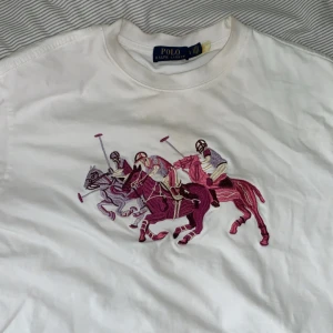 Vit Polo Ralph Lauren t-shirt  - Vit t-shirt från Polo Ralph Lauren med färgglad broderad polospelare på bröstet i rosa och lila toner. Klassisk rund hals och korta ärmar. Tillverkad i mjuk bomull, perfekt för en clean och sportig look.