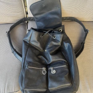 Svart ryggsäck i skinn med dragkedjor - Snygg svart ryggsäck i skinn med två framfickor som har silvriga dragkedjor och coola detaljer. Väskan har snörning upptill och ett lock med magnetknäppning. Perfekt för dig som vill ha en stilren och praktisk väska till vardagen.